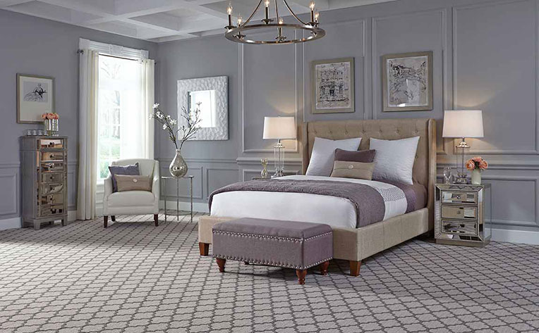 Large open bedroom decorated with a bed on a tan frame, a purple ottoman, lamps on nightstands, a white chair, and a candle light-themed chandelier on earth-toned patterned carpet.